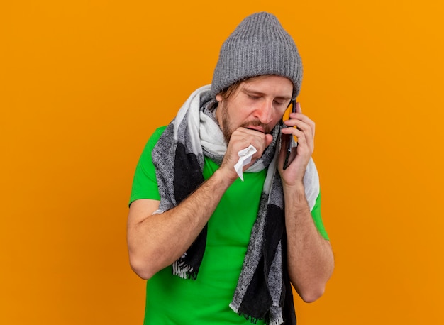 Gratis foto zwakke jonge knappe slavische zieke man met winter hoed en sjaal praten aan de telefoon houden servet hoesten vuist houden in de buurt van mond met gesloten ogen geïsoleerd op een oranje achtergrond met kopie ruimte