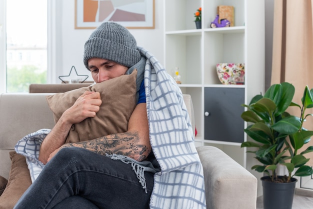 Zwakke jonge zieke man met sjaal en wintermuts zittend met gekruiste benen op de bank in de woonkamer gewikkeld in deken knuffelend kussen naar beneden kijkend