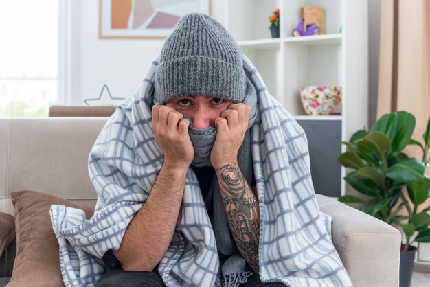 Zwakke jonge zieke man met sjaal en wintermuts zittend op de bank in de woonkamer gewikkeld in deken kijkend naar camera die mond bedekt met sjaal