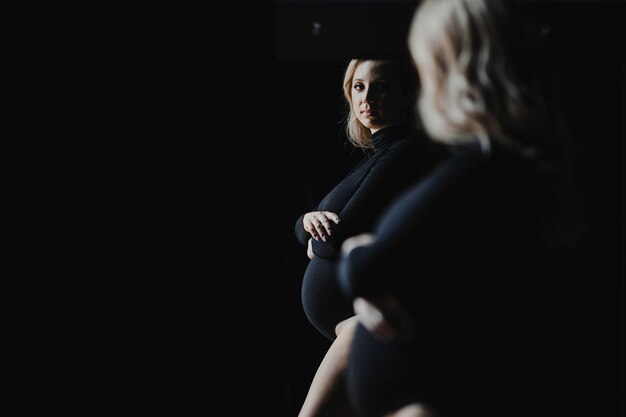 Zwangere blonde vrouw in een zwarte bodysuit staat in de buurt van een spiegel en kijkt naar haar spiegelbeeld