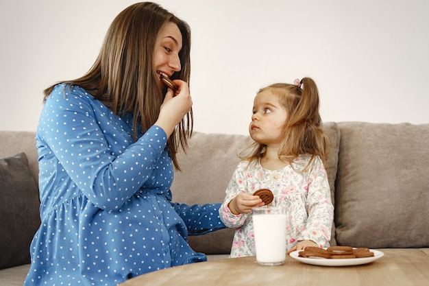 Zwangere moeder in een jurk. Meisje drinkt melk. Moeder en dochter genieten van koekjes.