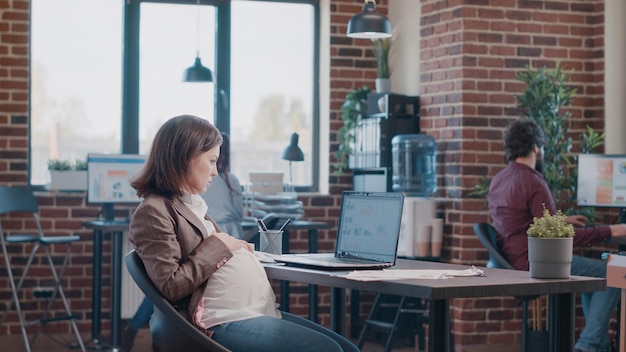 Zwangere vrouw die laptop gebruikt om aan zakelijk project op kantoor te werken. ondernemer verwacht kind dat met computer werkt voor ontwikkelings- en marketingstrategie. werknemer maakt presentatie