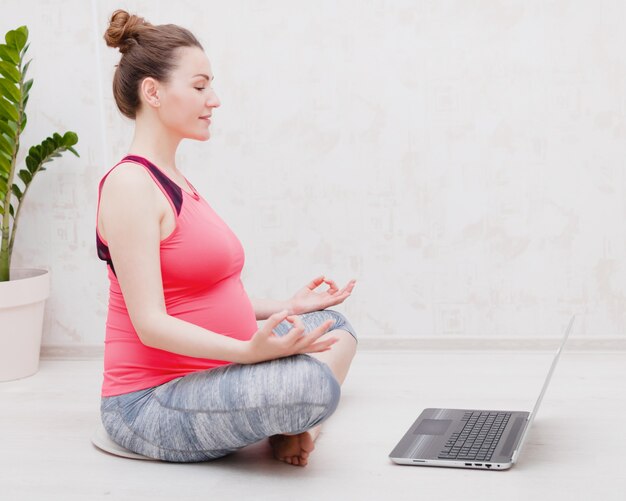 Zwangere vrouw maakt yoga praktijk online klas thuis, kijkt naar de laptop in yoga pose