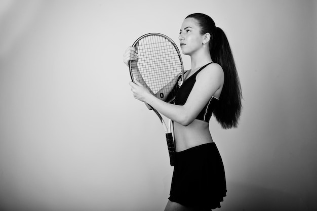 Zwart-wit portret van een mooie jonge vrouw die in sportkleding een tennisracket vasthoudt terwijl ze tegen een witte achtergrond staat