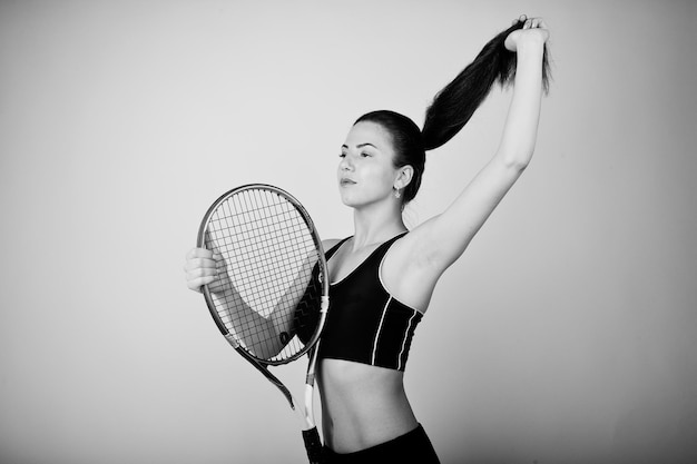 Zwart-wit portret van een mooie jonge vrouw die in sportkleding een tennisracket vasthoudt terwijl ze tegen een witte achtergrond staat