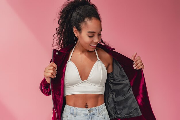 zwarte afro-amerikaanse vrouw in stijlvolle outfitjeans en fluwelen paars jasje, witte top op roze