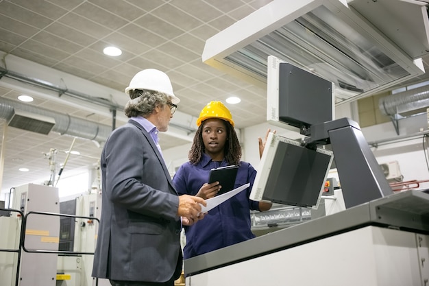 Zwarte fabrieksarbeider en haar mannelijke baas staan op industriële machine en praten