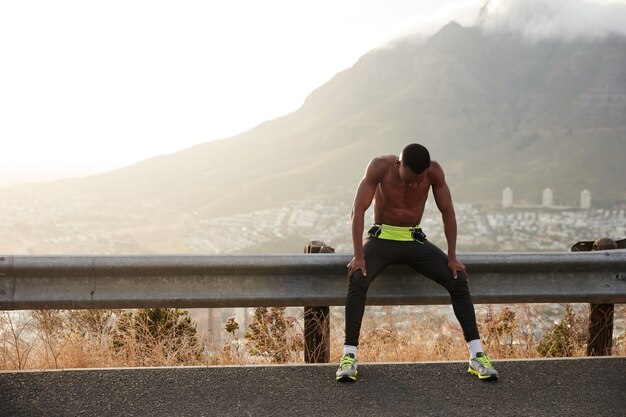 Zwarte gemotiveerde sportman voelt zich uitgeput na een actieve fitnesstraining, geniet van een vrije levensstijl, rust uit na het joggen in de ochtend, houdt de blik omlaag, heeft kort haar, gespierd atletisch lichaam, oefeningen buiten