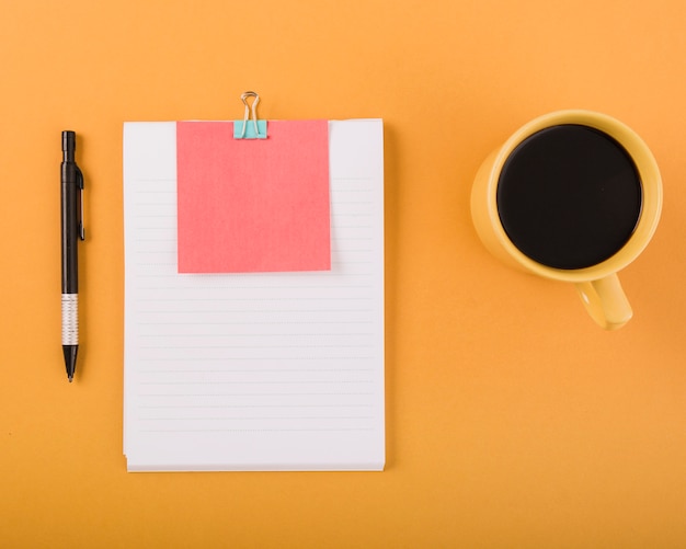 Zwarte koffie; pen en blanco papier met zelfklevende notitie op oranje achtergrond