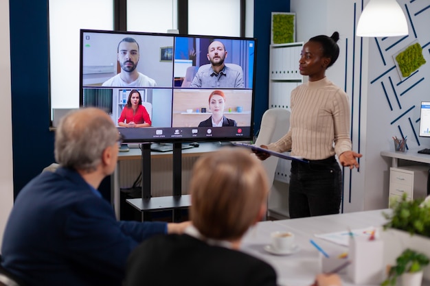 Zwarte managervrouw die op afstand met collega's praat tijdens een videogesprek op het tv-scherm en nieuwe zakenpartners voorstelt