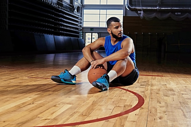 Zwarte professionele basketbalspeler zit op een vloer in een speelhal.