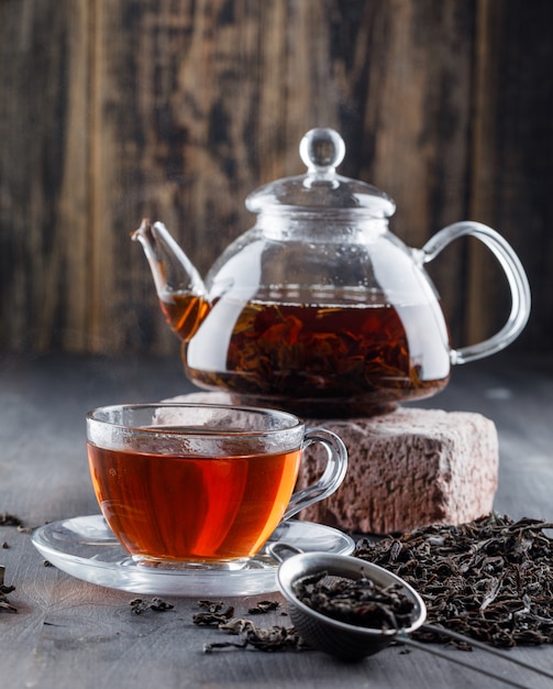 Zwarte thee in theepot en kopje met droge thee, bakstenen zijaanzicht op een houten oppervlak