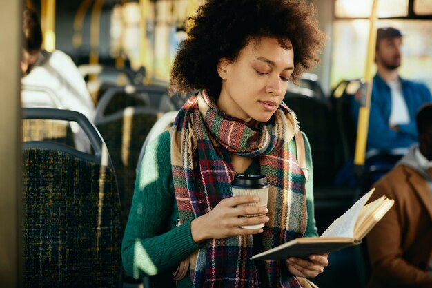 Zwarte vrouw leest een roman en drinkt afhaalkoffie terwijl ze met de bus reist