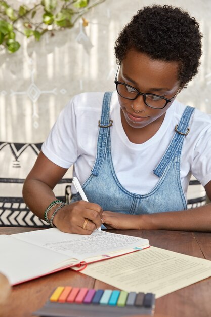 Zwarte vrouw met jongensachtig kapsel, schrijft in notitieboekje met pen, probeert cursuswerk te voltooien