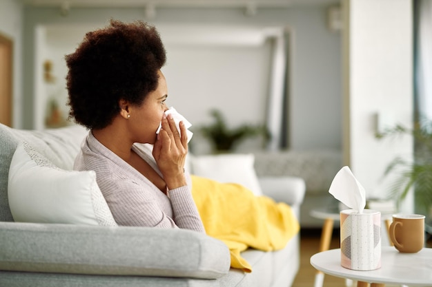 Gratis foto zwarte vrouw voelt zich misselijk en snuit haar neus in een tissue terwijl ze in de woonkamer rust