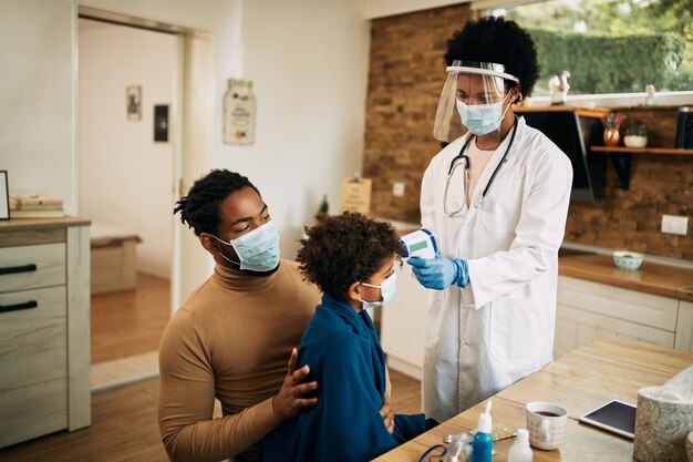 Zwarte vrouwelijke kinderarts die de temperatuur van de jongen meet tijdens een huisbezoek vanwege de COVID19-pandemie