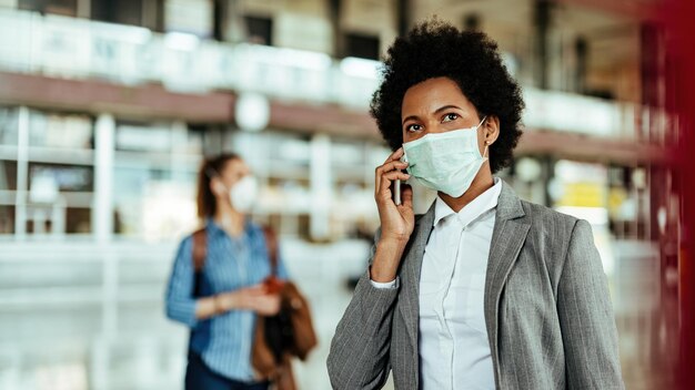 Zwarte vrouwelijke passagier die op een smartphone praat terwijl ze een gezichtsmasker draagt op de luchthaven tijdens een virusepidemie