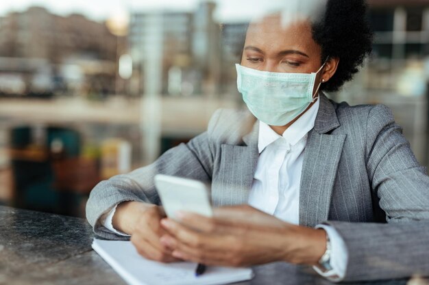 Zwarte zakenvrouw die een beschermend masker op haar gezicht draagt tijdens het gebruik van een smartphone en het lezen van een sms-bericht Het uitzicht is door het glas