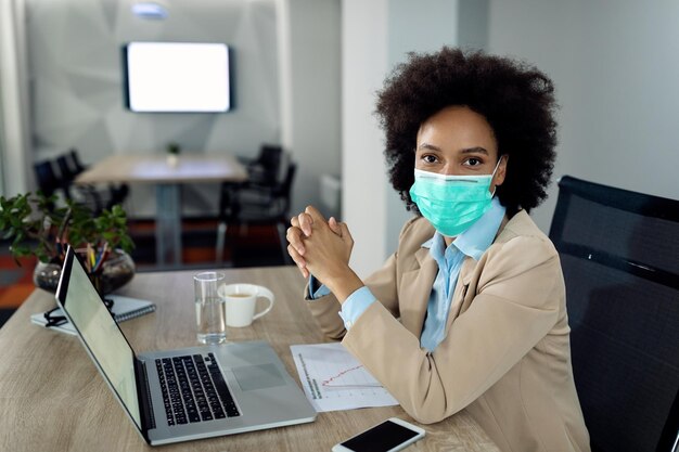 Zwarte zakenvrouw met beschermend gezichtsmasker die op laptop op kantoor werkt